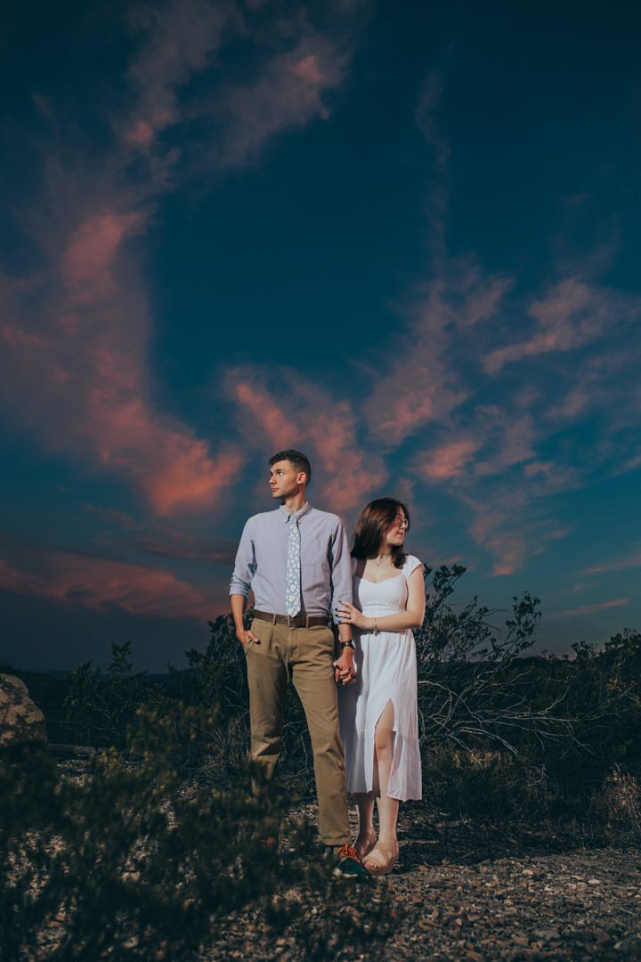 Newly engaged couple holding hands and standing together in the Big Bend National Park TX desert scrubland at dusk, dramatic deep blue and pink twilight sky with wispy clouds overhead as they pose for a romantic portrait in the Chihuahuan Desert, Big Bend surprise proposal post-engagement portrait photography Big Bend National Park TX surprise proposal photographer