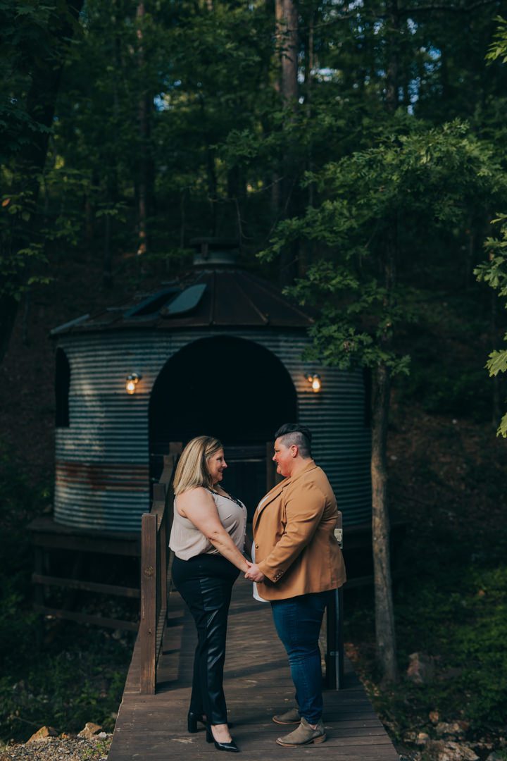 LGBTQ plus-size same-sex couple standing face to face holding hands on wooden bridge deck with rustic barrel cabin lit by warm lanterns behind them at Broken Bow Oklahoma, romantic dusk engagement portrait after queer surprise proposal, Broken Bow OK same-sex engagement photographer