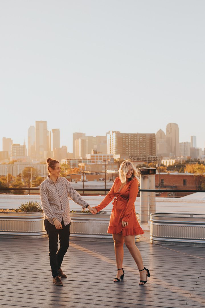 Same-sex couple spinning and dancing on a rooftop deck in Dallas Texas at golden hour with the full Dallas skyline behind them after a surprise proposal, playful LGBTQ queer couple engagement portrait, Dallas TX rooftop same-sex proposal photographer