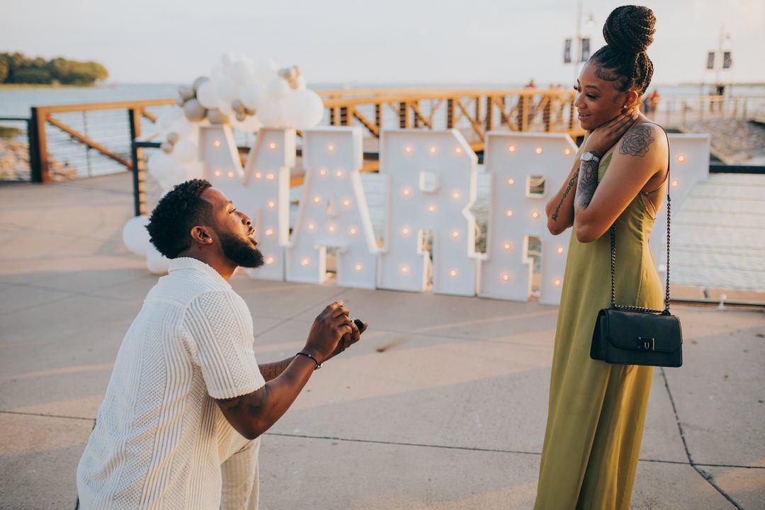 Black man down on one knee holding out an engagement ring to his girlfriend in front of glowing marquee letters and white balloon arrangements in Rockwall TX, Black woman smiles with emotion and touches her neck as she looks down at him during a surprise proposal on the Lake Ray Hubbard waterfront at golden hour, surprise proposal ring moment photography Rockwall TX surprise proposal photographer Dallas TX