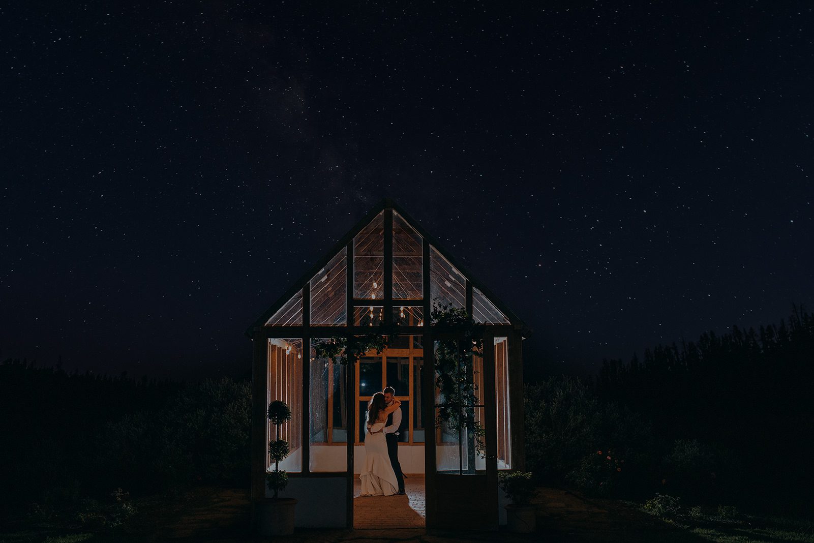 Moody wedding portrait of a bride and groom inside a glass greenhouse chapel at night, illuminated with warm lighting, surrounded by nature and a starry sky. Romantic editorial wedding photography for modern couples in Dallas and beyond.